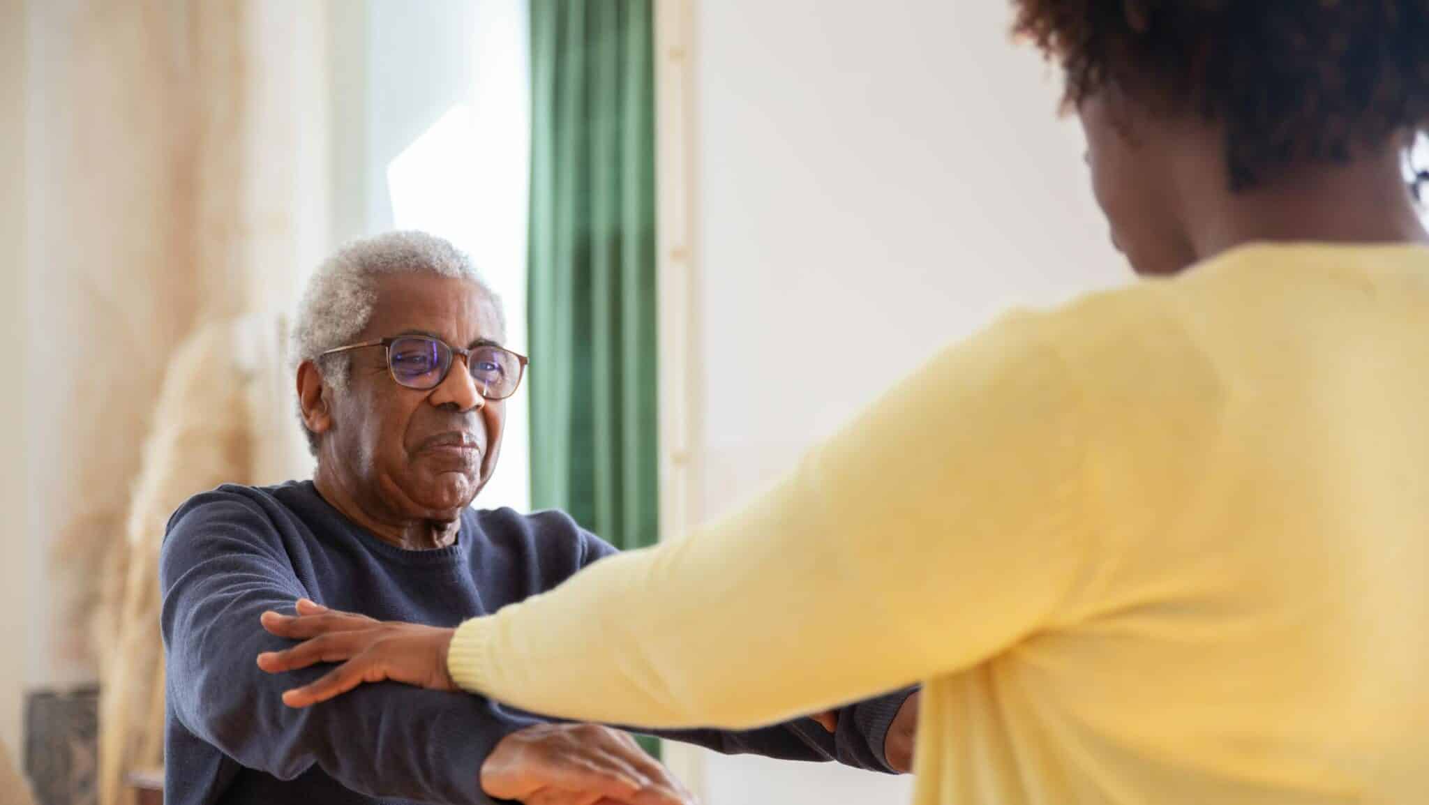 An elderly man with a caregiver assisting him in exercise at home.